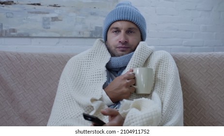 Sick Man In Warm Blanket And Hat Sitting On Couch With Cup Of Tea And Watching Tv