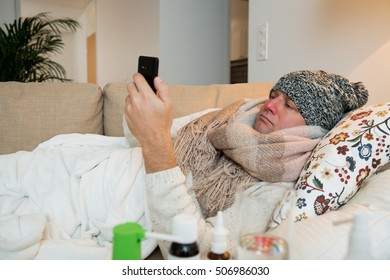 Sick Man Wearing Scarf Lying On Couch At Home Under A Blanket Checking Temperature. Browsing With Smart Phone. Living Room With Table Full Of Medicine And Pills. Man With Running Nose And Cough. 