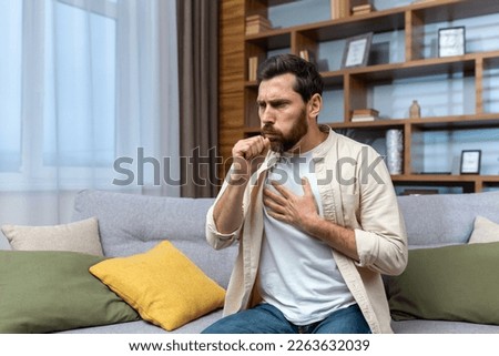 Sick mature man alone at home sitting on sofa coughing holding hands to chest in living room.