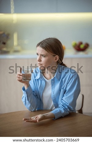 Sick middle-aged female sitting at table with pills, painkillers, antibiotics on palm, hold glass of water, looking at window, going to take treatment. Depressed girl suffering from headache, pain