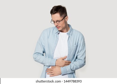 Sick Millennial Guy In Glasses Suffering From Stomach Ache, Holding Belly, Feeling Discomfort. Upset Man With Abdomen Pain, Gastritis Symptom, Indigestion Problem, Isolated On Grey Studio Background.