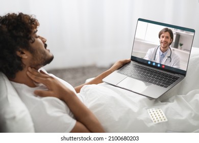 Sick millennial hindu guy having video chat with his male doctor, sitting in bed, using laptop, touching his throat, suffering from angina or tonsillitis, side view. Remote medical care concept