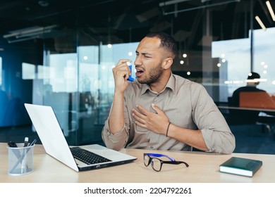 Sick Office Worker With Asthma Having Trouble Breathing, Man Using Inhaler To Relieve Breathing, Hispanic Man Working Inside Modern Office Building, Mixed Race Man With Laptop.