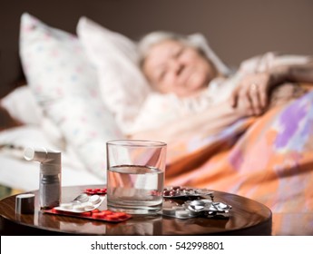 Sick Old Woman Lying In Bed At Home. Focus On Medicine And Glass Of Water In Foreground