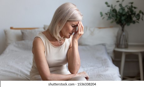 Sick Older Mature Woman Sit On Bed Feel Pain Dizziness Coping With Morning Headache Concept, Upset Tired Middle Aged Adult Woman Touching Sore Head Suffer From Terrible Migraine Mental Problem