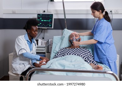 Sick Patient Lying In Bed While Woman Nurse Putting Oxygen Mask Monitoring Respiratory Illness. Physician Black Medic Writing Sickness Treatment Working In Hospital Ward During Recovery Appointment