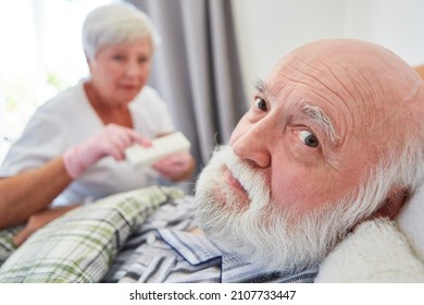 Sick Senior In Bed In Nursing Home And Geriatric Nurse With Medication Box In The Background