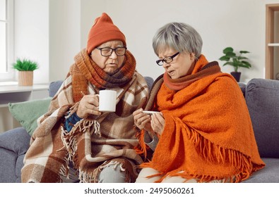 Sick senior couple covered in blankets sitting on sofa at home with seasonal flu or cold. Elderly woman looking at thermometer checking temperature feeling unhealthy with influenza. - Powered by Shutterstock