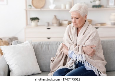 Sick Senior Lady Sitting On Couch Covered With Blanket, Freezing Shivering Due To High Temperature, Suffering From Coronavirus At Home