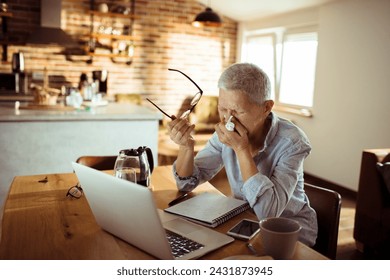 Sick senior woman blowing nose in paper tissue while using laptop at home - Powered by Shutterstock