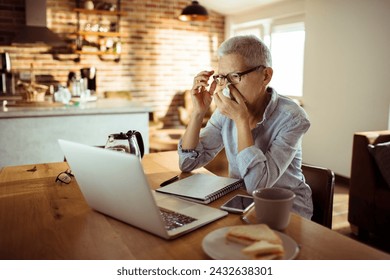 Sick senior woman blowing nose in paper tissue while using laptop at home - Powered by Shutterstock