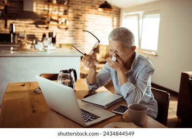 Sick senior woman blowing nose in paper tissue while using laptop at home - Powered by Shutterstock