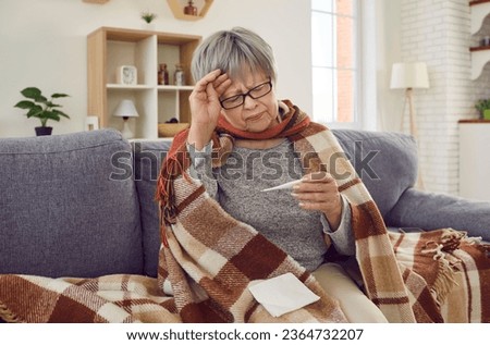 Sick senior woman checking her temperature wrapped in a blanket on the sofa suffering from headache. Elderly person with seasonal flu or cold feel unhealthy with influenza at home.