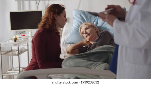 Sick Senior Woman Patient Lying In Hospital Bed With Adult Daughter Sitting Near And Listening To Doctor. Practitioner Discussing Diagnosis With Sick Mature Female And Daughter Visitor In Hospital