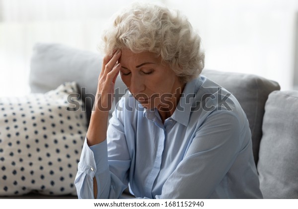 Sick senior woman sit on sofa at home feel\
distressed suffer from migraine or headache, ill mature female rest\
on couch touch head have dizziness from high blood pressure,\
elderly healthcare concept