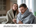 Sick shivering couple of man and woman wrapped in blanket. Male measuring fever on couch, battling flu symptoms and bad health at home. Family sick leave, virus, pandemic illness at home, quarantine