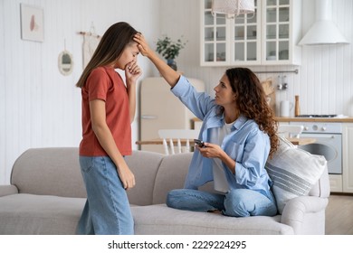 Sick suffering teenage girl approaches mother to complain about feeling unwell and coughing. Caring mysterious woman measures daughter's temperature puts hand to forehead sits on sofa in apartment - Shutterstock ID 2229224295