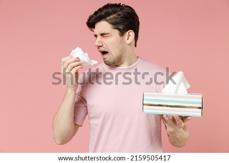 Sick unhealthy ill allergic man has red watery eyes runny stuffy sore nose suffer from allergy trigger symptoms hay fever hold paper napkin handkerchief isolated on pastel pink color background studio