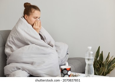 Sick unhealthy woman with hair bun sitting on sofa wrapped in blanket, having illness, suffering runny nose and sneezing, posing in room at home, needs treatment.