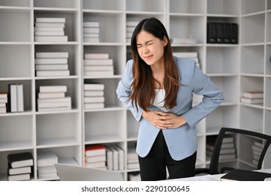 Sick and unwell millennial Asian businesswoman suffering from stomachache or abdominal pain in her office. - Shutterstock ID 2290100645