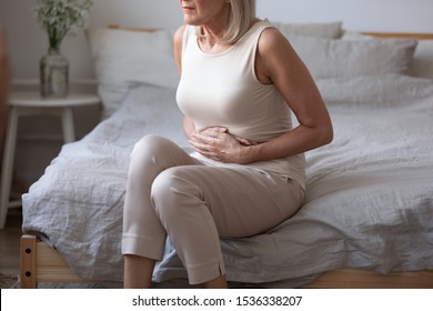 Sick upset middle aged lady hold belly suffer from abdomen ache concept, mature old adult woman feel morning pain hurt in stomach abdominal gastritis pancreatitis diarrhea problem symptom sit on bed