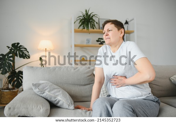 Sick upset middle aged lady hold belly suffer from\
abdomen ache concept, mature old adult woman feel morning pain hurt\
in stomach abdominal gastritis pancreatitis diarrhea problem\
symptom sit on bed