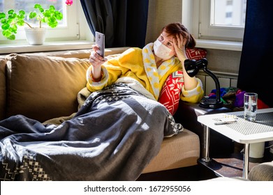 Sick Woman In A Bathrobe With A Medical Mask Lies In The Home Interior On The Couch. She Called A Doctor By Phone