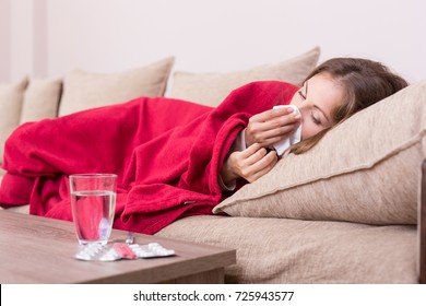 Sick woman covered with a blanket lying in bed with high fever and a flu, blowing her nose. Pills and glass of water on the table