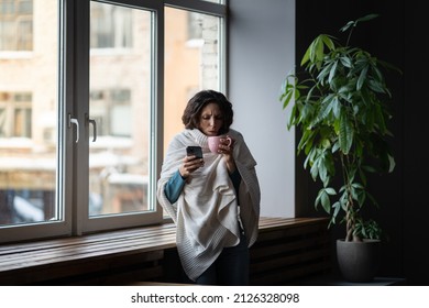 Sick Woman Having COVID Infection Symptoms Typing Message, Sms Or Sick Day Email To Manager, Contacting Boss And Inform Of Sick Day By Phone While Standing Wrapped In Plaid Near Window At Home