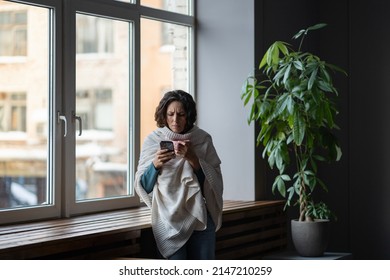 Sick Woman Having COVID Infection Symptoms Typing Message, Sms Or Sick Day Email To Manager, Contacting Boss And Inform Of Sick Day By Phone While Standing Wrapped In Plaid Near Window At Home