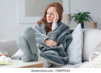 Sick woman with a headache sitting on a sofa at home wrapped in grey blanket