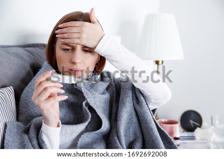 Sick woman lying in bed in self isolation at coronavirus covid-19 quarantine, measuring temperature with thermometer touching forehead, having fever. Infected patient at home medical self treatment. 