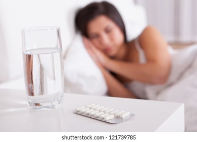 Sick Woman Resting In Bed With Focus To A Glass Of Water And A Blister Pack Of Tablets And Medication Lying On The Table Alongside Her Bed