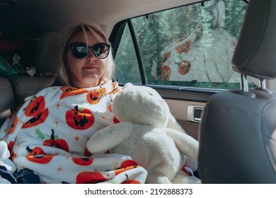 Sick Woman Rides In The Backseat Of A SUV Car While On A Road Trip, Covered In A Blanket, Holding A Stuffed Animal Rabbit Bunny