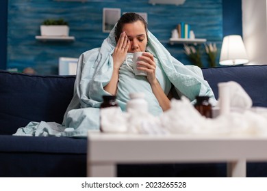 Sick Woman Sitting At Home In Blanket With Hot Tea Suffering From Fever Temperature Infection Flu Headache Cold. Young Person Drinking Tea, With Seasonal Virus Symptoms Feeling Unwell