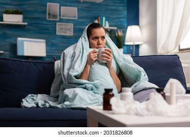 Sick Woman Sitting At Home In Blanket With Hot Tea Suffering From Fever Temperature Infection Flu Headache Cold. Young Person Drinking Tea, With Seasonal Virus Symptoms Feeling Unwell