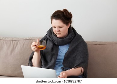 A sick woman is sitting at home with hot tea, communicating with friends and family online, with a laptop on her lap. - Powered by Shutterstock