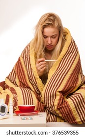 Sick Woman Sitting On Bed Wrapped In A Blanket Feeling Ill, Has Flu And Fever, Looking In Thermometer.