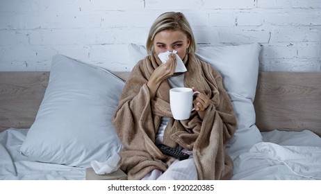 Sick Woman Sneezing Into Paper Napkin While Watching Tv In Bedroom With Cup Of Tea