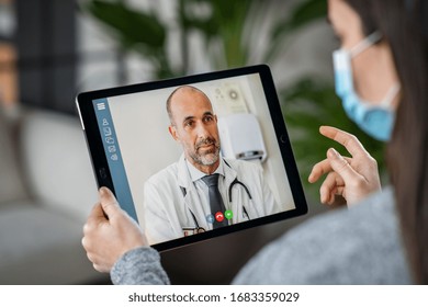 Sick woman wearing protective mask during video call with mature doctor. Back view of patient with surgical face mask talking during conference call with her physician, staying at home in quarantine. 
