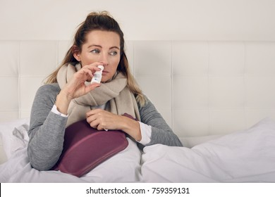 Sick Woman Wrapped In A Warm Scarf And Clutching A Hot Water Bottle Sitting Using Nasal Spray With A Miserable Expression In A Seasonal Healthcare Or Flu Concept