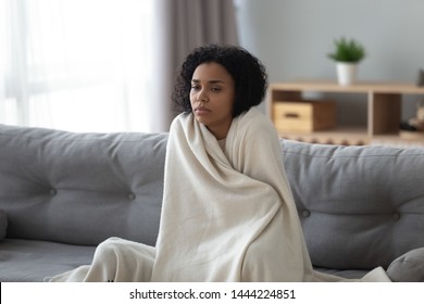 Sick young african woman feeling cold covered with blanket sit on sofa, ill black girl shivering freezing warming at home wrapped with plaid, no central heating problem, fever temperature flu concept