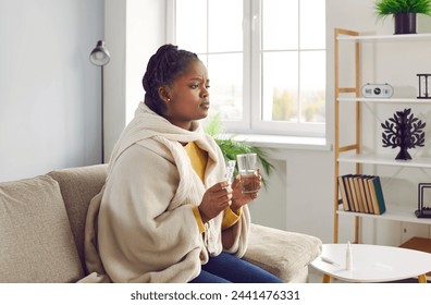 Sick young africanamerican woman feels unwell and is going to drink medicine, washed down with a glass of water.Girl is sitting at home warmly dressed and wrapped in a warm blanket.Health care concept