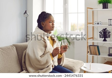 Sick young africanamerican woman feels unwell and is going to drink medicine, washed down with a glass of water.Girl is sitting at home warmly dressed and wrapped in a warm blanket.Health care concept