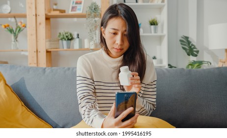 Sick Young Asian Woman Hold Medicine Sit On Couch Video Call With Phone Consult With Doctor At Home. Girl Take Medicine After Doctor Order, Quarantine At Home, Social Distancing Coronavirus Concept.