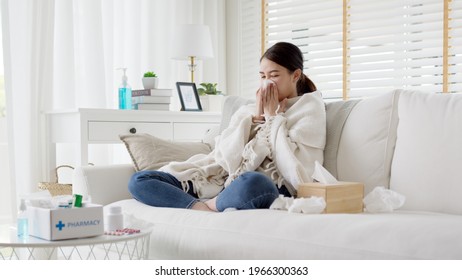 Sick young asian woman sitting under the blanket on sofa and sneeze with tissue paper at home. Female blowing nose, coughing or sneezing in tissue at home, suffering from flu. Cold and fever concept - Shutterstock ID 1966300363