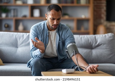 Sick Young Black Man Measuring Arterial Tension While Sitting On Couch At Home, African American Guy Using Electronic Sphygmomanometer, Checking Blood Pressure With Upper Arm Monitor In Living Room