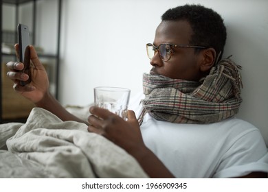 Sick Young Black Man Staying Home Because Of Respiratory Disease Texting Message To Boss On Cell Phone, Taking Medication With Water, Lying In Bed Wearing Warm Scarf Around Neck. Health And Technology