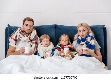 Sick Young Family With Cups Of Hot Drink Sitting In Bed And Looking At Camera