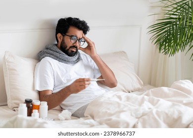 Sick Young Indian Man Looking At Thermometer While Sitting In Bed At Home, Ill Eastern Guy Checking Body Temperature And Touching Aching Head, Suffering Seasonal Flu Symptoms, Copy Space